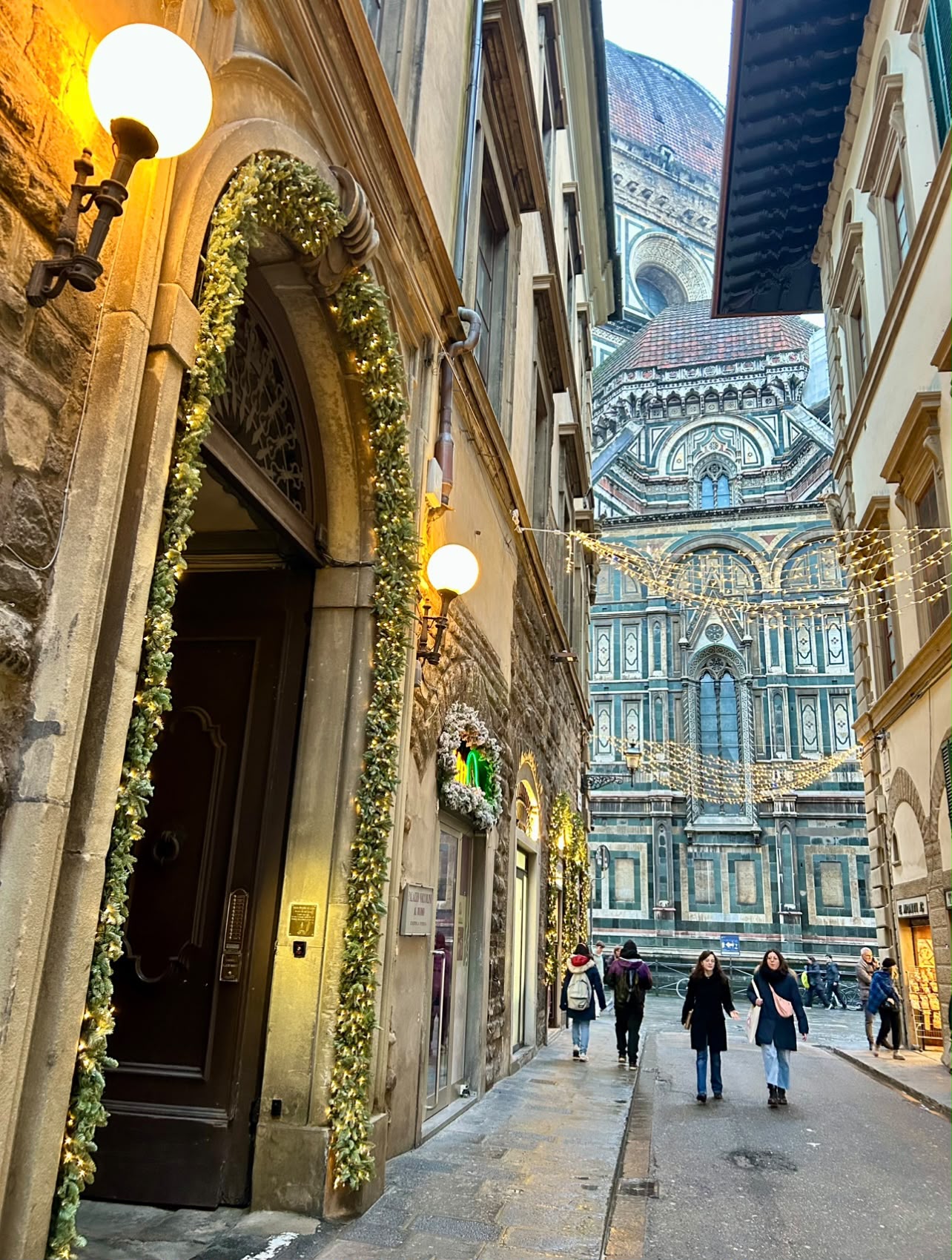 Via dei Servi illuminata la sera, con il portone adornato di ghirlande e il Duomo in fondo